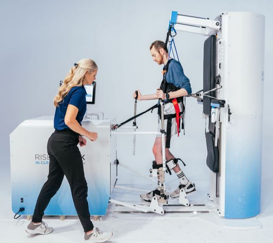 Client using a Rise & Walk rehabilitation device with therapist assistance during gait training session, supported by harness and leg braces in a clinical setting.